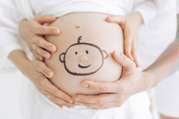 Couple parents touch pregnant belly by their hands with drawn smiling baby face on it . Concept of love and care during pregnancy, expecting and born baby.
